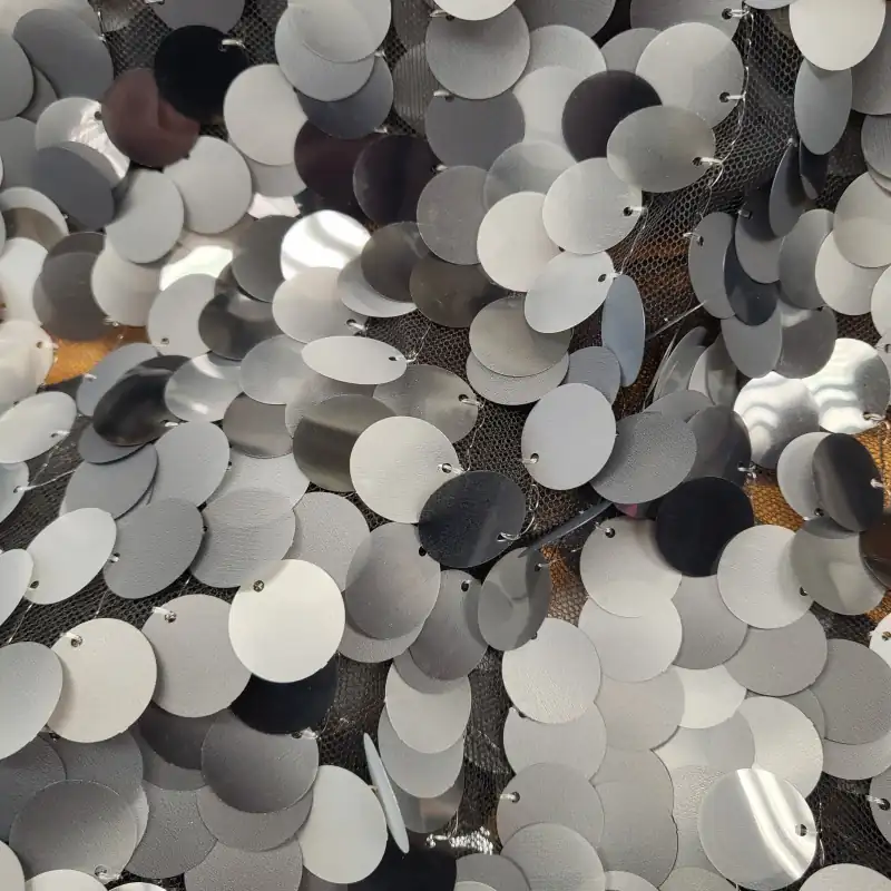 Κοντινό πλάνο από επικαλυπτόμενες στρογγυλές ασημί και γκρι παγιέτες ραμμένες πάνω σε μαύρο πλεγμένο ύφασμα, που αντανακλούν το φως και δημιουργούν μια λαμπερή, με υφή επιφάνεια - ιδανική για υφάσματα με παγιέτες. Υφάσματα Ιχτιάρογλου