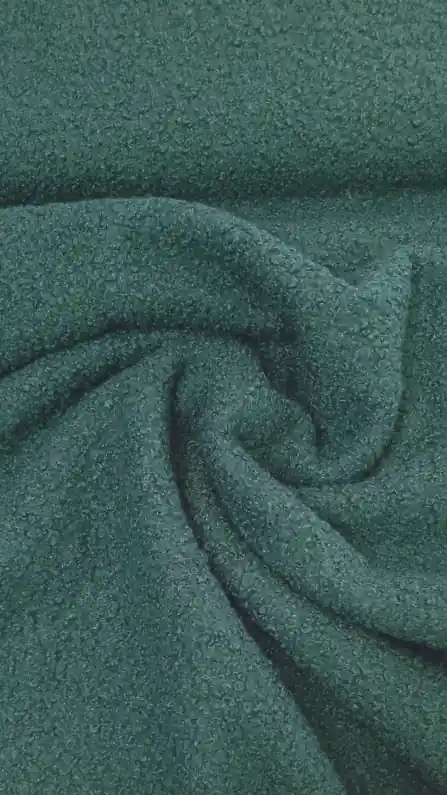Κοντινό πλάνο του Μπουκλέ ελαστικό 13-044 σε σκούρο πράσινο χρώμα, με μαλακή, χνουδωτή επιφάνεια. Οι απαλές πτυχώσεις και το χαλαρό μοτίβο στροβιλισμού αναδεικνύουν τη ζεστή υφή του, ιδανική για μπουκλέ ζεστά ρούχα. Υφάσματα Ιχτιάρογλου