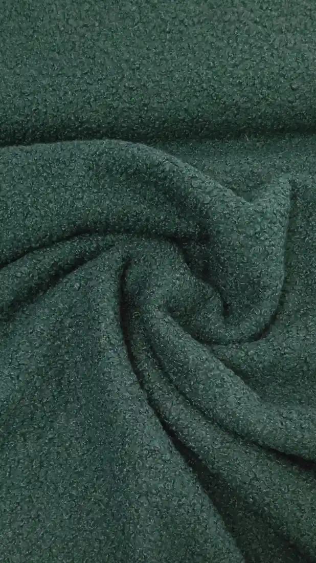 Ένα κοντινό πλάνο σκούρου πράσινου, υφασμάτινου υφάσματος με μαλακή, σγουρή επιφάνεια -όπως το Μπουκλέ ελαστικό 13-044- τοποθετημένο σε ένα απαλό μοτίβο στροβιλισμού, ιδανικό για μπουκλέ ζεστά ρούχα. Υφάσματα Ιχτιάρογλου