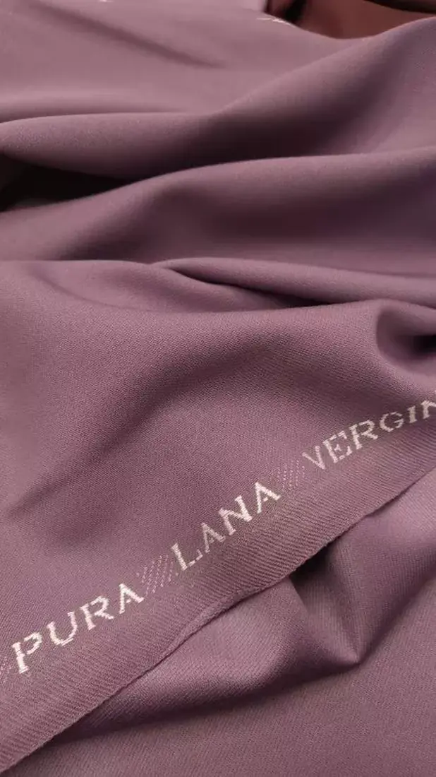 Κοντινό πλάνο από μαλακό, μοβ ύφασμα για φόρεμα με τις λέξεις PURA LANA VERGIN υφασμένες με λευκό χρώμα κατά μήκος της άκρης. Το ιταλικό μαλλί charmelaine φαίνεται λείο και ελαφρώς διπλωμένο. Υφάσματα Ιχτιάρογλου