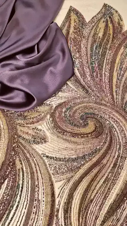 Ένα κομμάτι από λαμπερό Τούλι κεντημένο 24061, με περίτεχνη χάντρα με πέρλα παγιέτα σε χρυσά, χάλκινα και μωβ σχέδια paisley, βρίσκεται δίπλα σε ένα διπλωμένο, λείο μωβ σατέν ύφασμα. Υφάσματα Ιχτιάρογλου