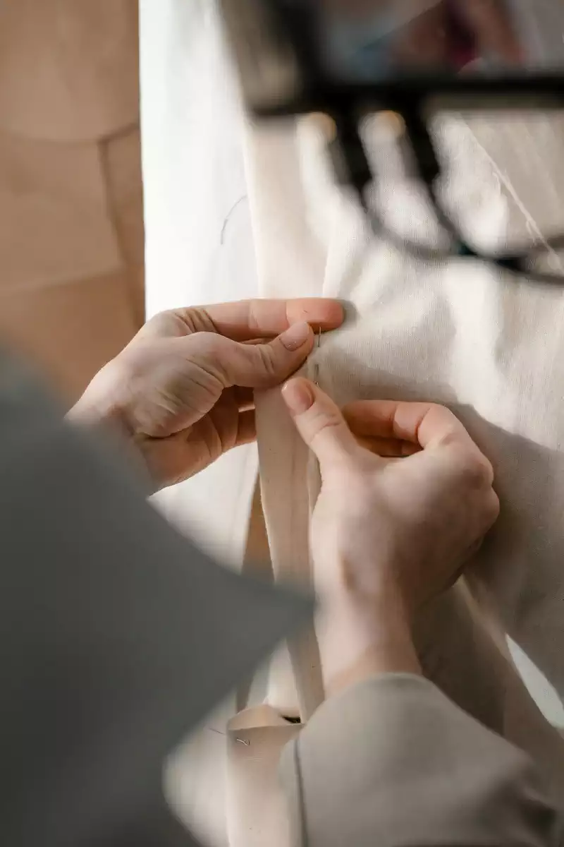 Κοντινό πλάνο των χεριών μισς σχεδιάστριας που καρφιτσώνει ή ράβει ένα κομμάτι από ανοιχτόχρωμα, υψηλής ποιότητας υφάσματα, με θολό φόντο και απαλό φυσικό φωτισμό. Υφάσματα Ιχτιάρογλου