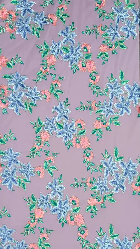 Ένα λεβάντα ύφασμα με κεντημένο floral μοτίβο, στο οποίο απεικονίζονται συστάδες από μπλε και ροζ λουλούδια με πράσινα φύλλα, διάσπαρτα ομοιόμορφα σε όλη την επιφάνεια. Υφάσματα Ιχτιάρογλου